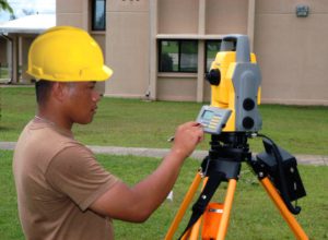 A black man wearing a yellow construction hat and he is adjusting a theodolite on a beautiful summer day