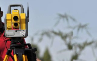 Image of a black man standing behind a surveyors tripod with a theodolite, he is ging instructions to the other surveyor to move right