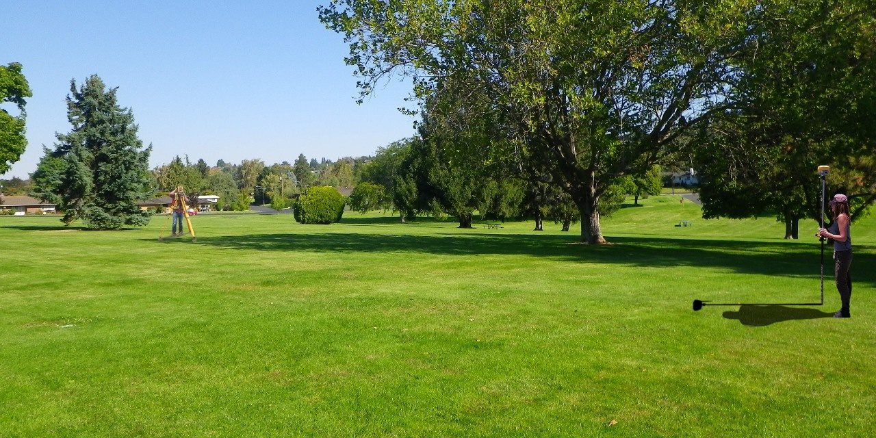 open grass field with large trees on the border on a bright sunny day with a woman and man conducting a survey
