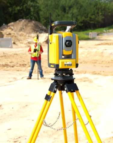 A survey expert on a construction site holding a clipboard while another expert surveyor looks through a theodolite