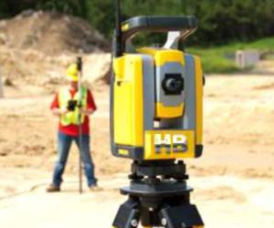 image of a residential survey expert holding a leveling rod or prism pole with a blurred out theodolite in the foreground pointing at the pole. It looks like a very sunny and hot summer day. 