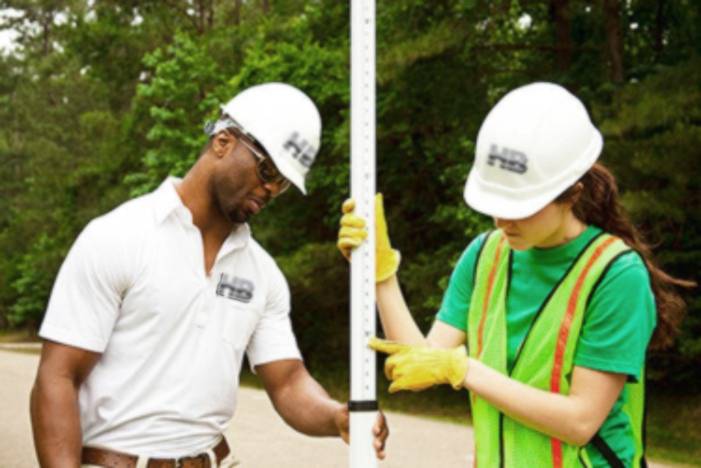 an image of a black man of color and a caucasian woman looking at a leveling rod or prism pole. It looks to be a very sunny summer day.