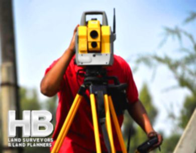 An image of a man of color using a theodolite and he is performing a Property Survey in Frederick, Maryland on a beautiful sunny summer day, there is not a single cloud in the sky and it looks hot.
