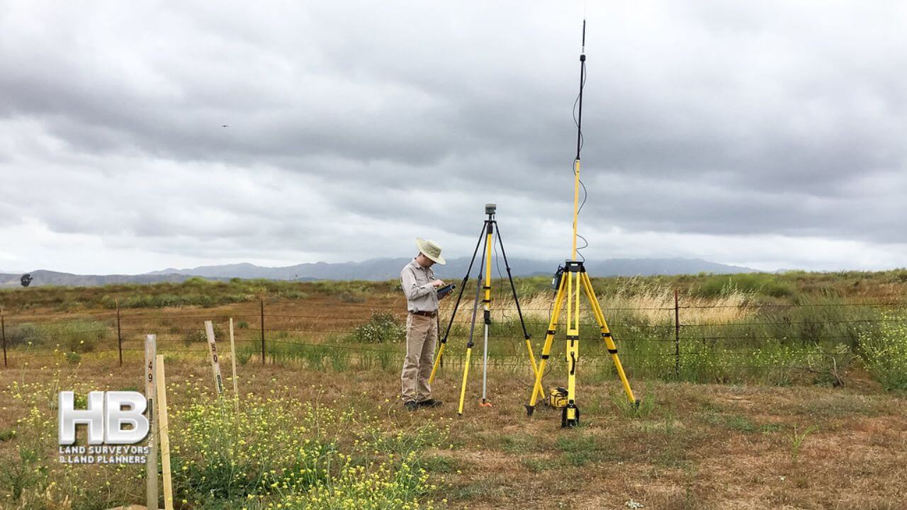 A man stands in a field looking at a tablet and configuring settings for two communicating pieces of modern land surveying quipment.