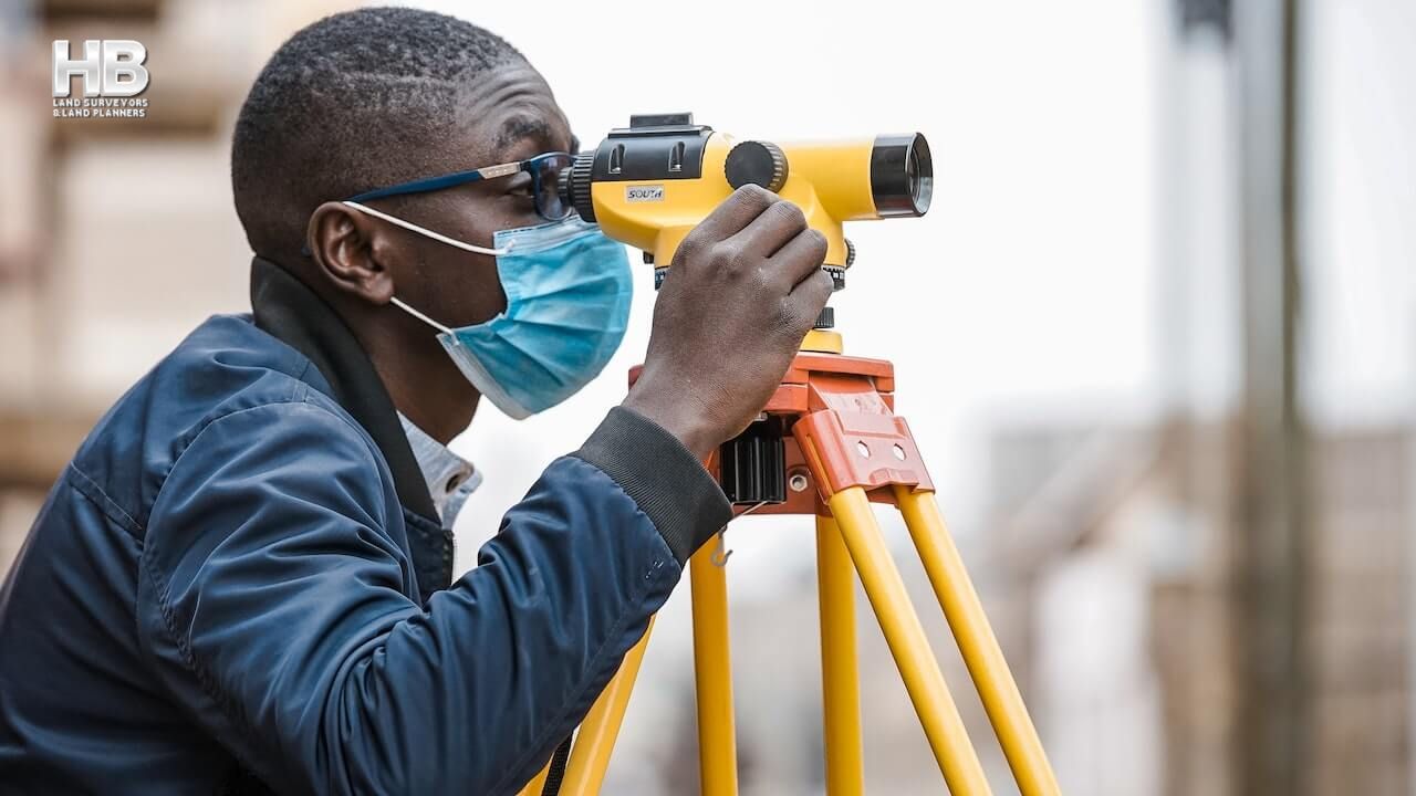 An African American man wearing dark glasses and a cloth face mask looks through a piece of yellow equipment to perform topographical land surveys.
