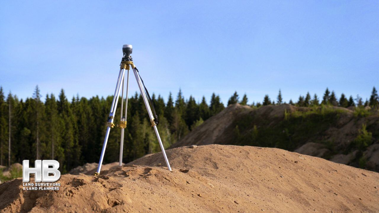 A piece of equipment for land surveys set-up on a large mound of brown coarse earth with a forest of green pine or fir trees in the distance.