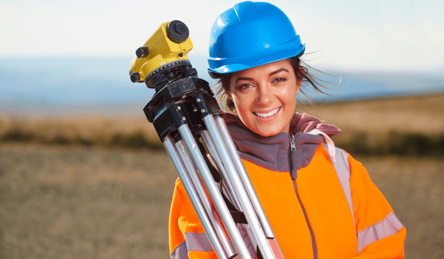 One young, attractive woman smiling in a rural scenic area only looking directly at camera holding a surveyors level measurement tool wearing reflective surveying industrial clothing and protective headwear.