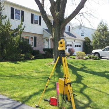 an image of a average American Home 2 story, attached garage a big tree in the front yard, the sun is shining and there is a theodolite in the fore ground