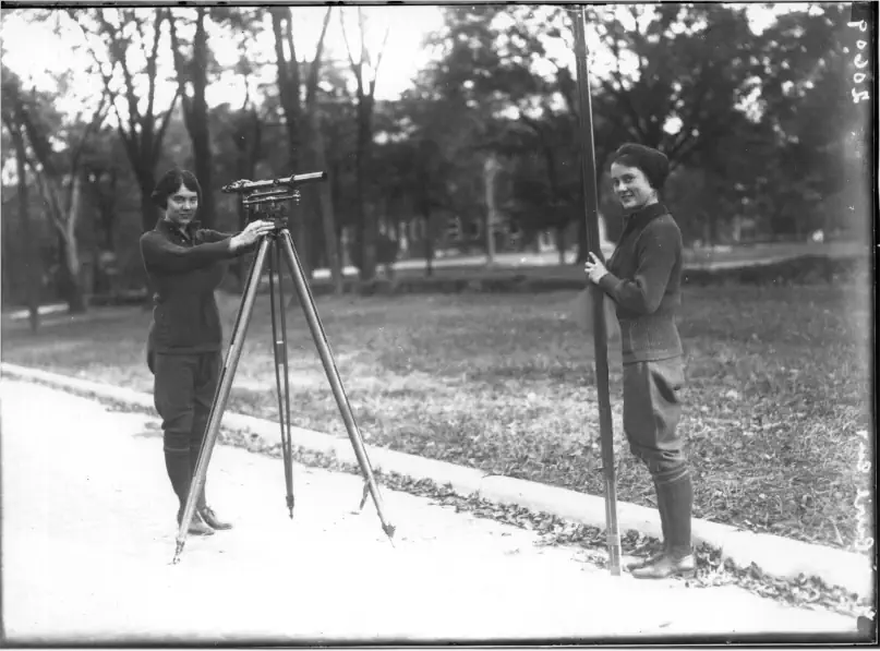an image from circa 1920's black and white 2 woman surveyors looking at the camera and smiling, the look very happy, in their boots, bloomers and black sweaters, its a very cool image.